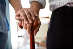 The hand of an elderly patient holding a cane with the hand of a medical professional resting on top for comfort.