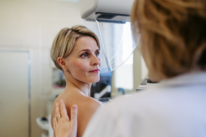 A woman with short blonde hair who is about to undergo a mammogram while the technologist has her hand on her back to gently position her.