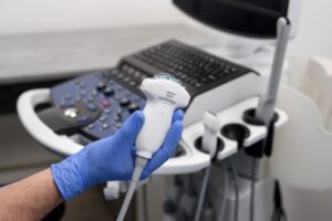 A hand with a blue glove holds a tool for ultrasound and there is a table with a keyboard and nobs in the background. 