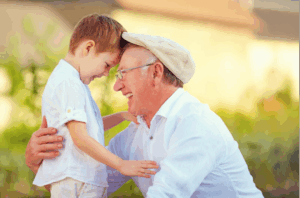 A grandpa wearing a white shirt smiling with his arm around his grandson who is also smiling whil their foreheads touch.