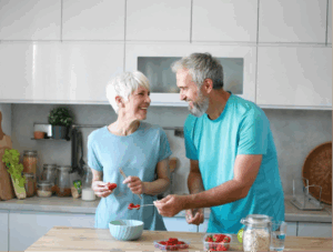 An older couple weating blue shirts smiling at each other in the kitchen while they make breakfast.