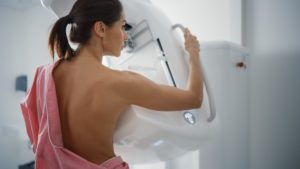 A woman with a pink gown draped over one shoulder facing away and holding onto a handle on a mammogram machine