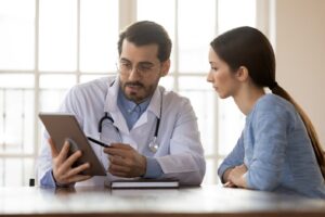 A Doctor Wearing a Lab Coat and Stethoscope Points to a Tablet and a Woman Sitting Next to Him Looks at the Tablet