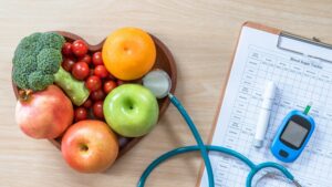 Fruits and Vegetables in Heart-Shaped Bowl with a Stethoscope in it and a Clipboard and Blood Sugar Testing Device Next to It