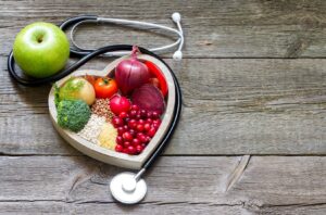 A heart surrounding heart healthy fruit, vegetables, and grains surrounded by a stethoscope on a wood plank floor.