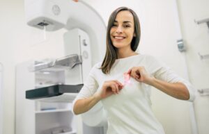 Woman smiling in front of a mammography machine while holding a pink breast cancer ribbon. 
