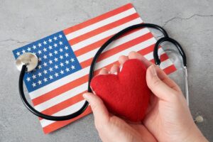 A small American flag on a stone surface beneath a stethoscope and hands holding a red felt heart. 