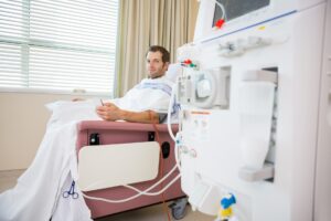 A man in a chair under a blanket with a tube running from under the blanket to a dialysis machine in the foreground
