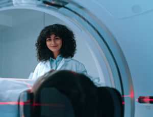 Smiling medical professional standing near an imaging machine with a patient pictured from behind undergoing a scan.