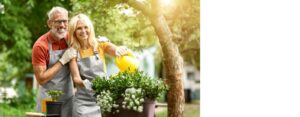 An older couple wearing matching aprons, smiling together while watering their plants outside.