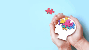 Hands protecting white paper head silhouette with colorful puzzle pieces inside, red piece above