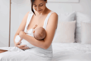 A woman sitting on a bed while smiling down at her baby and breastfeeding the baby.