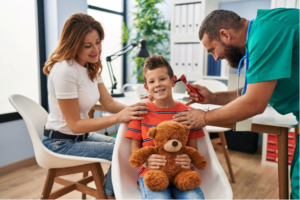 A little boy holding a teddy bear while his mom comforts him and a doctor looks in his ear. 
